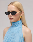 A woman with straight blonde hair models For Art’s Sake® Abstraction I bold temple sunglasses, gold earrings, and a light blue pleated sleeveless top against a simple, light background.