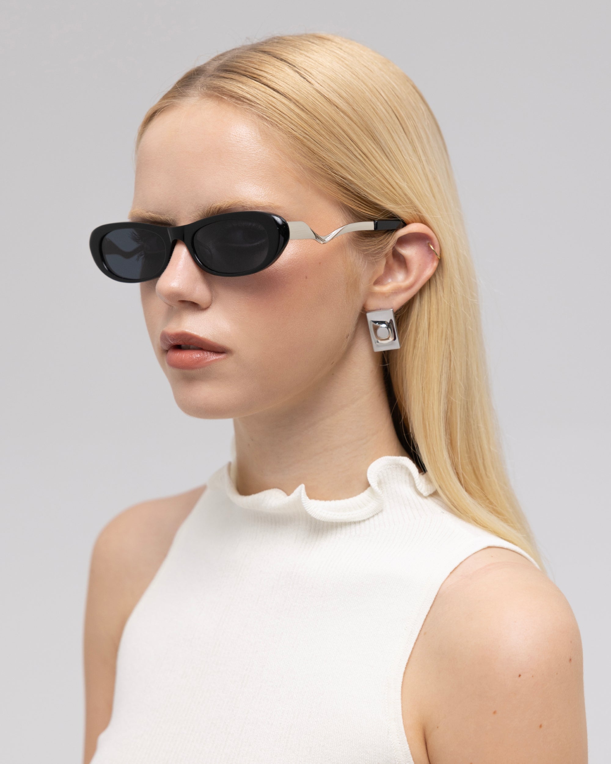 A woman with long blonde hair models For Art's Sake® Abstraction I sculptural sunglasses, geometric silver earrings, and a sleeveless white ruffled top against a light gray background.