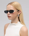 A woman with long blonde hair models For Art's Sake® Abstraction I sculptural sunglasses, geometric silver earrings, and a sleeveless white ruffled top against a light gray background.