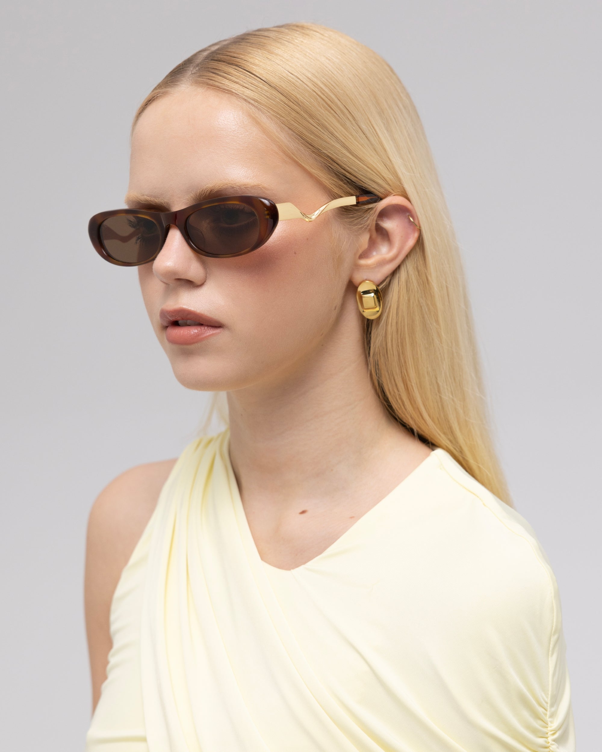 A woman with long blonde hair wears For Art's Sake® Abstraction I oval sculptural sunglasses, a pale yellow sleeveless top, and gold geometric earrings against a plain light background.