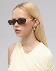 A woman with long blonde hair wears For Art's Sake® Abstraction I oval sculptural sunglasses, a pale yellow sleeveless top, and gold geometric earrings against a plain light background.