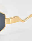 Close-up of For Art's Sake® Abstraction II statement eyewear with a gold geometric metal frame, dark lenses, and wavy sculpted temples, shown against a plain light background.