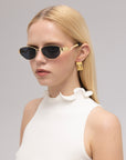 A woman with straight blonde hair wears For Art's Sake® Abstraction II black and gold statement sunglasses, geometric gold earrings, and a sleeveless white top with a ruffled collar against a plain background.