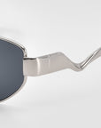 Close-up of For Art's Sake® Abstraction II sunglasses featuring a silver geometric metal frame, dark lenses, and distinctively curved, shiny temples against a plain light background.