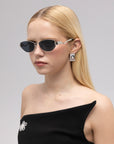 A woman with straight blonde hair wears For Art's Sake® Abstraction II sunglasses, geometric silver earrings, and a black off-shoulder dress with a jeweled brooch, posed against a plain light gray background.