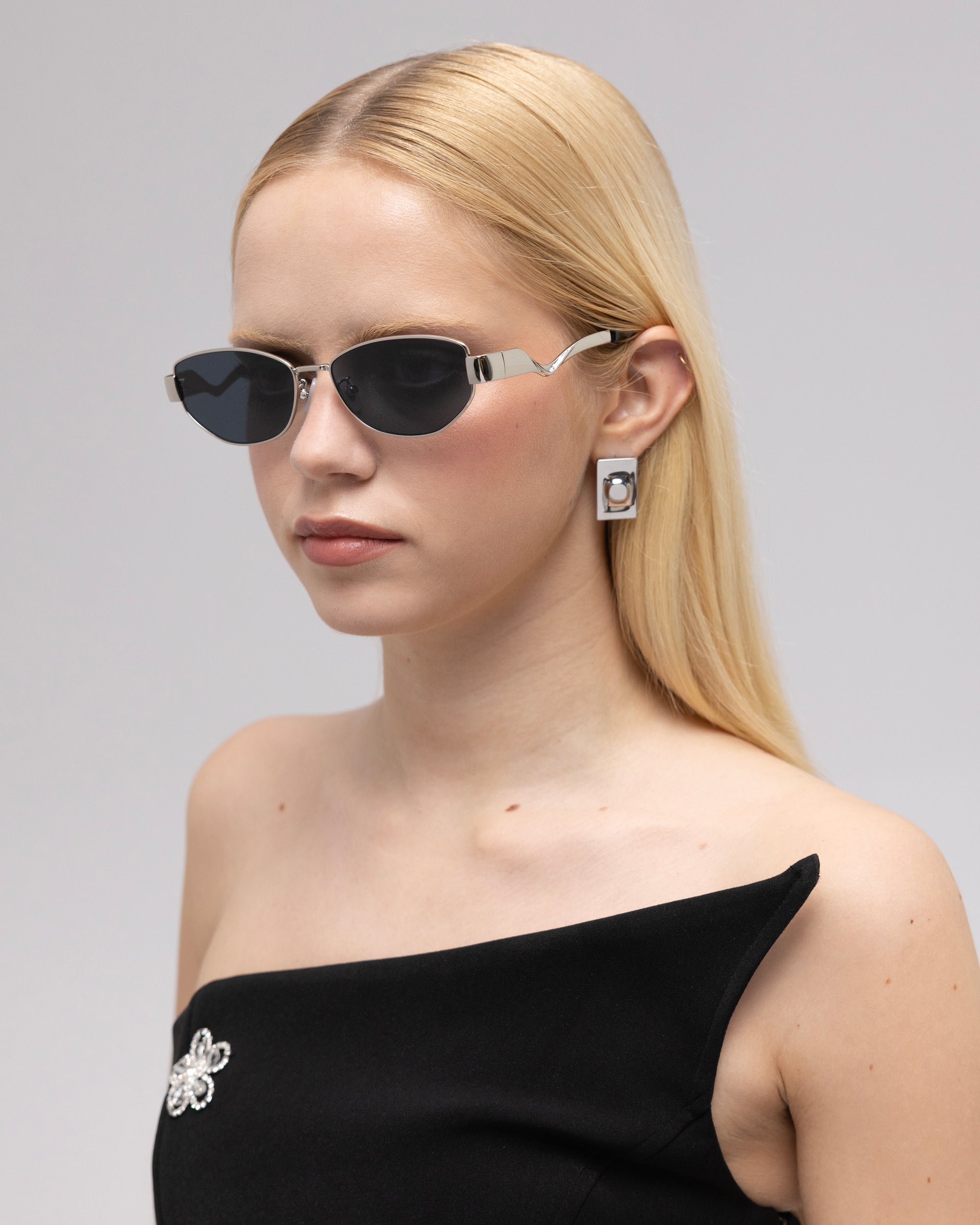 A woman with straight blonde hair models For Art's Sake® Abstraction II sculptural sunglasses, geometric earrings, and an off-shoulder black dress with a jeweled brooch, posing against a plain light background.