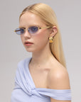 A woman with straight blonde hair poses against a neutral background wearing For Art's Sake® Mesa I sculptural sunglasses, gold square earrings, and a light blue off-shoulder top.