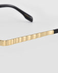 Close-up of the Mesa I temple arm by For Art’s Sake®, showing its gold metallic ripple texture and black hinge detail against a light gray background—a subtle expression of quiet luxury.
