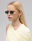 A woman with straight blonde hair wears For Art's Sake® Mesa I understated sunglasses, gold geometric earrings, and a pale yellow asymmetrical draped top with a subtle ripple texture against a neutral background.