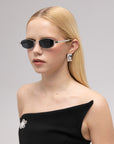 A woman with long blonde hair poses elegantly against a plain light background, wearing For Art's Sake® Mesa I sculptural sunglasses, rectangular photo earrings, and a one-shoulder black dress with a jeweled brooch.