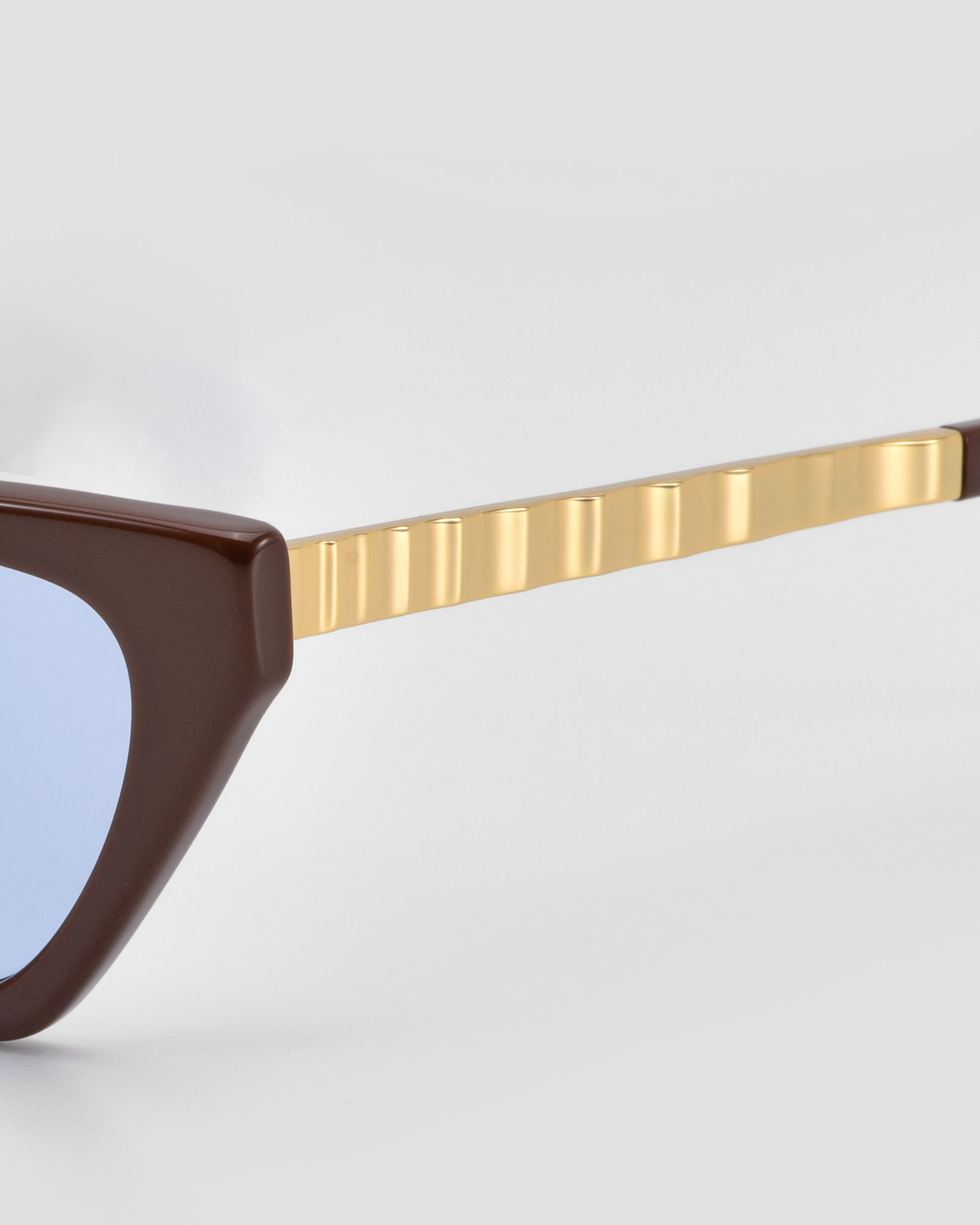 Close-up of For Art's Sake® Mesa II cat-eye sunglasses with a dark brown acetate frame, light blue lenses, and textured gold temple arms shown against a plain light gray background.