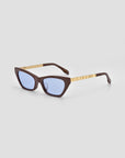 The Mesa II sunglasses by For Art's Sake® feature sculptural cat-eye shapes, dark brown frames, gold arms, and light blue tinted lenses, exuding quiet luxury against a plain light grey background.