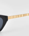 Close-up of the Mesa II sunglasses by For Art's Sake®, featuring a black frame, dark lenses, and a gold ribbed metal arm, set against a light gray background.