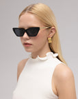 A woman with straight blonde hair wears For Art's Sake® Mesa II sculptural cat-eye sunglasses, gold earrings, and a sleeveless white top with a ruffled high neckline, posing against a plain light background.