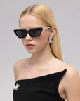 A woman with straight blonde hair models For Art's Sake® Mesa II luxury cat-eye sunglasses, geometric silver earrings, and an off-shoulder black dress with a jeweled brooch, set against a plain light background.