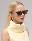 A woman with straight blonde hair models For Art's Sake® Mesa II brown acetate cat-eye sunglasses, gold earrings, and a sleeveless pale yellow top with a high collar against a plain light background.