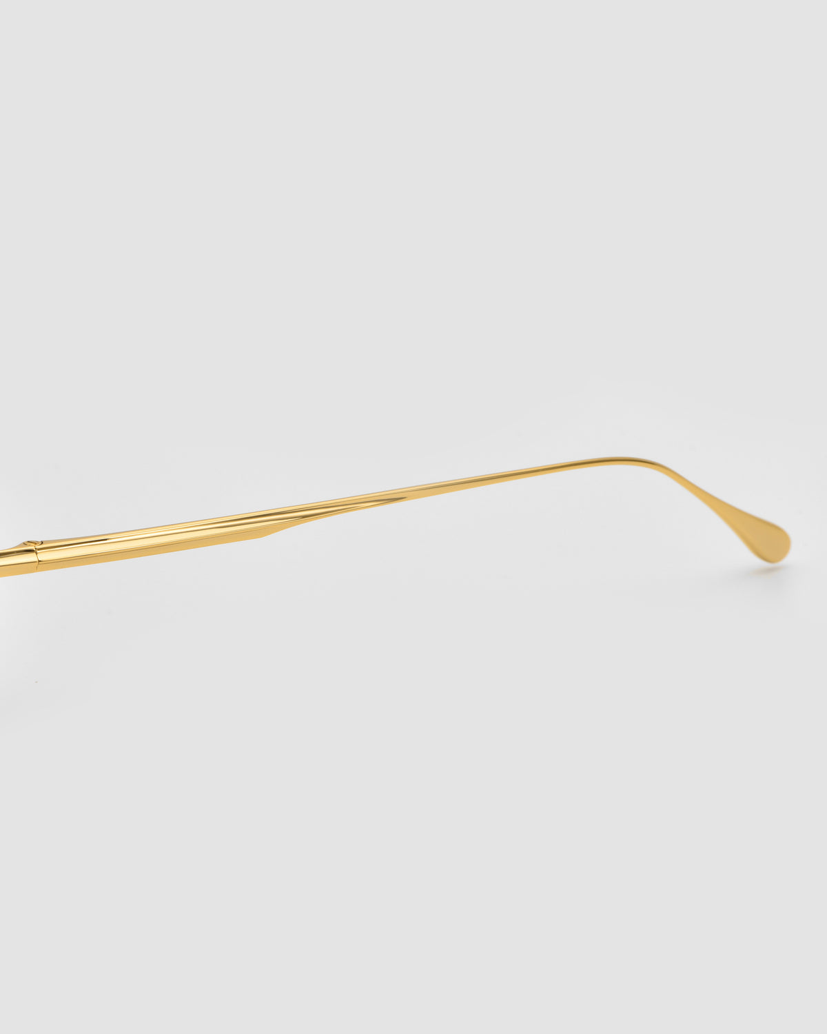 A single gold eyeglass arm from the For Art’s Sake® Sienna I sunglasses is displayed against a plain light gray background, extending horizontally with a gentle curve at the tip.