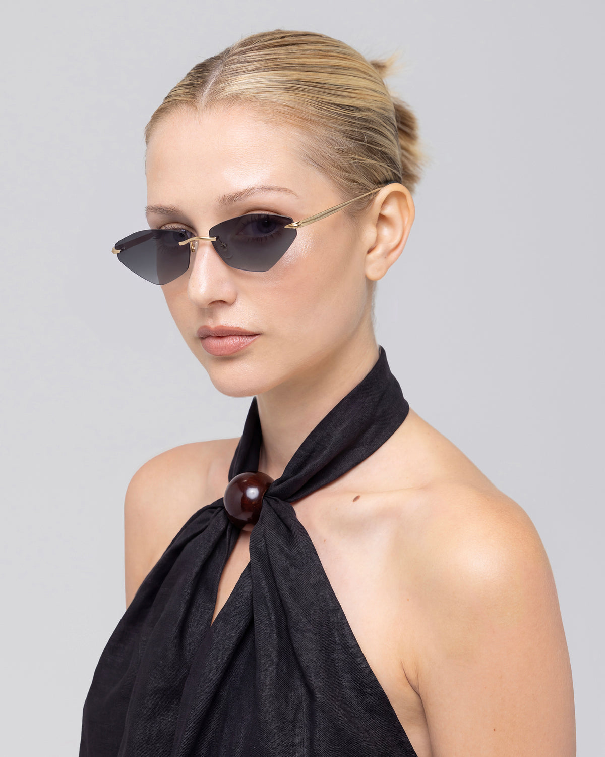 A woman with blonde hair in a bun is wearing For Art’s Sake® Sienna I dark rimless sunglasses featuring jade stone nose pads, along with a black halter top with a large round clasp, as she gazes slightly to the side against a light gray background.