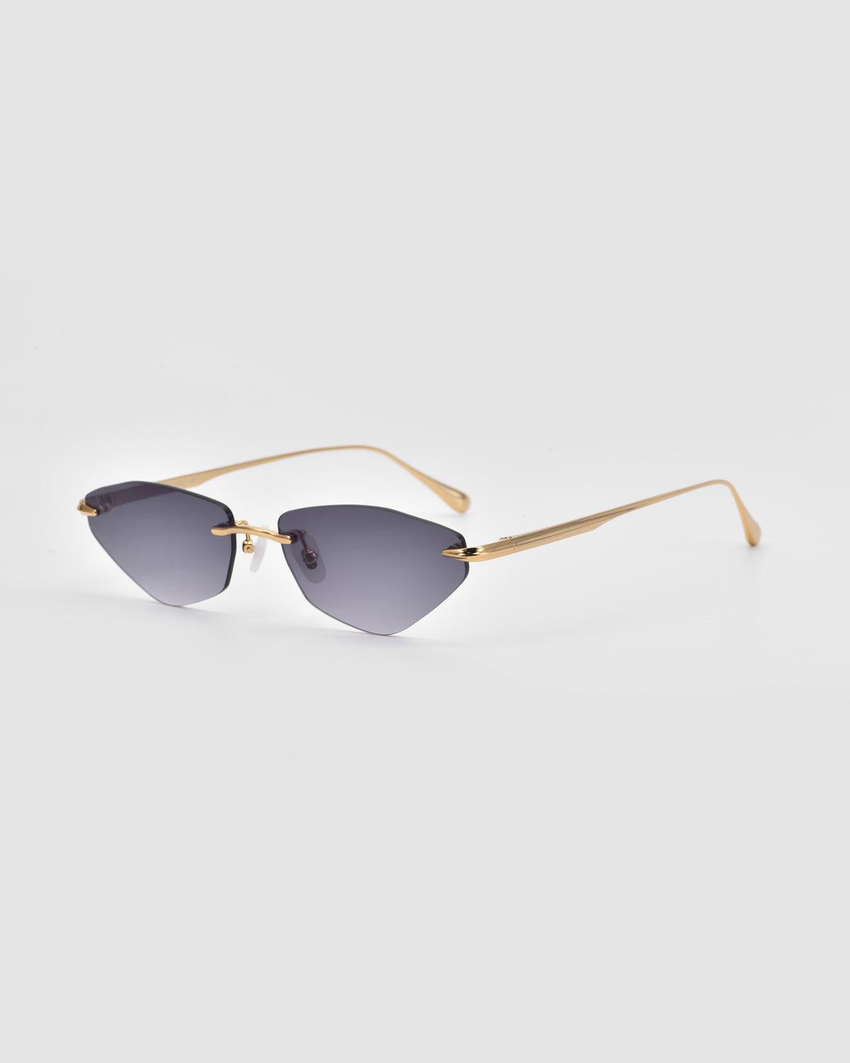 Sienna I sunglasses by For Art’s Sake® showcase gold frames and dark, cat-eye shaped lenses resting on a light gray background. Their minimal design is distinguished by thin arms, jade stone nose pads, and a sophisticated rimless silhouette.
