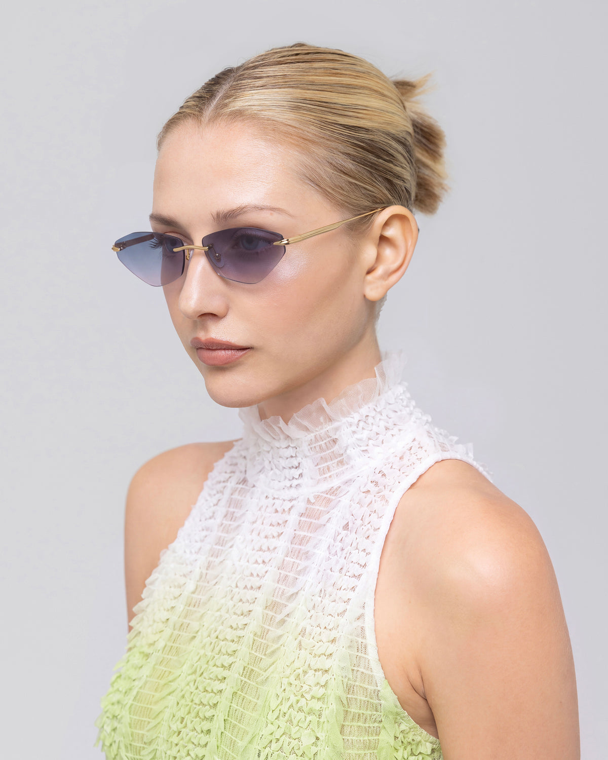 A woman with blonde hair in a bun models the For Art’s Sake® Sienna I sunglasses, featuring jade stone nose pads, paired with a sleeveless textured white and light green top as she poses against a plain light background.