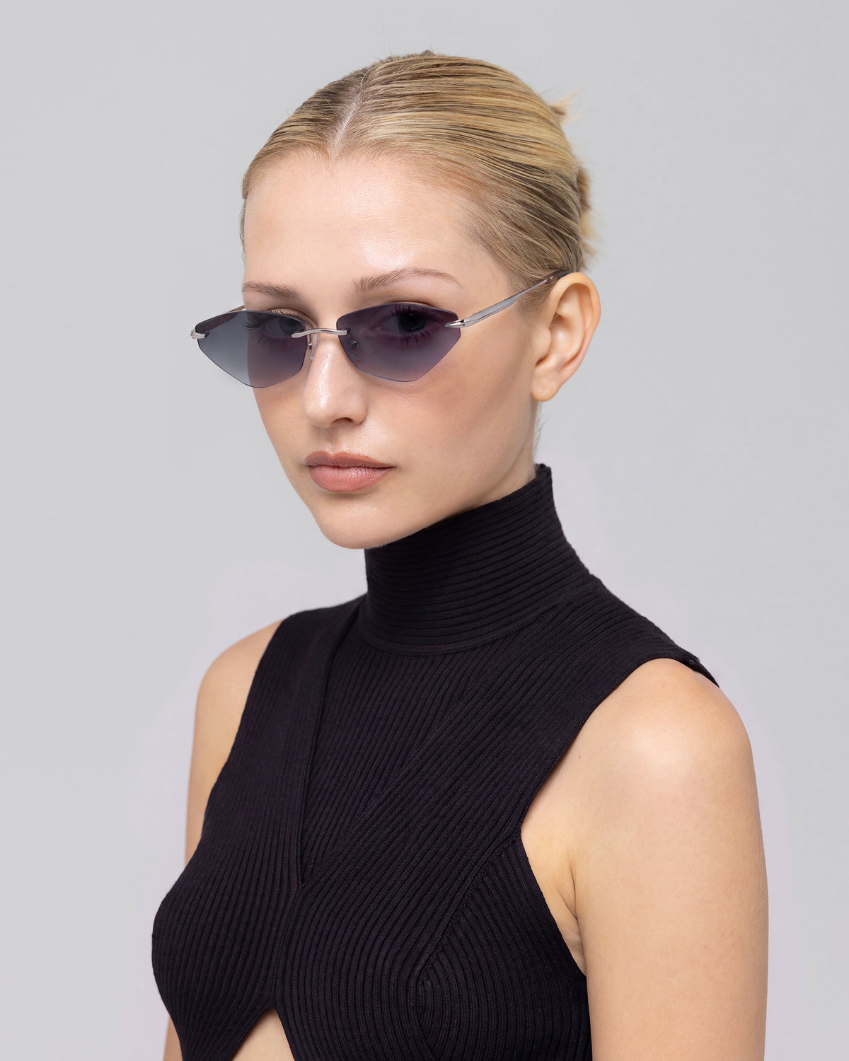A woman with slicked-back blonde hair wears For Art’s Sake® Sienna I rimless sunglasses featuring jade stone nose pads and a sleeveless black turtleneck top, gazing slightly away from the camera against a plain light gray background.