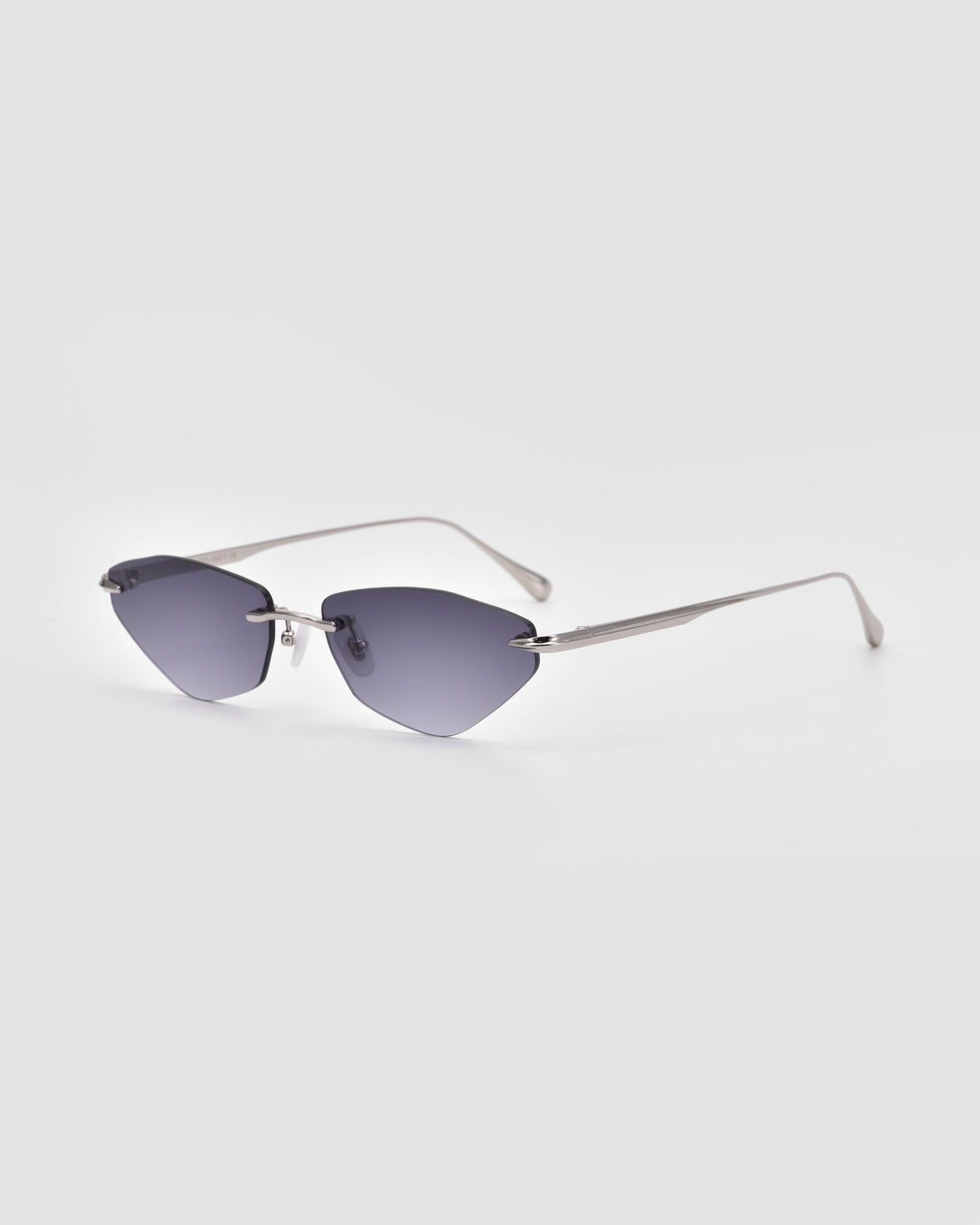 Sienna I by For Art’s Sake® features rimless sunglasses with angular, dark gray tinted lenses and slender silver metal arms, accented by jade stone nose pads, showcased against a plain light gray background.