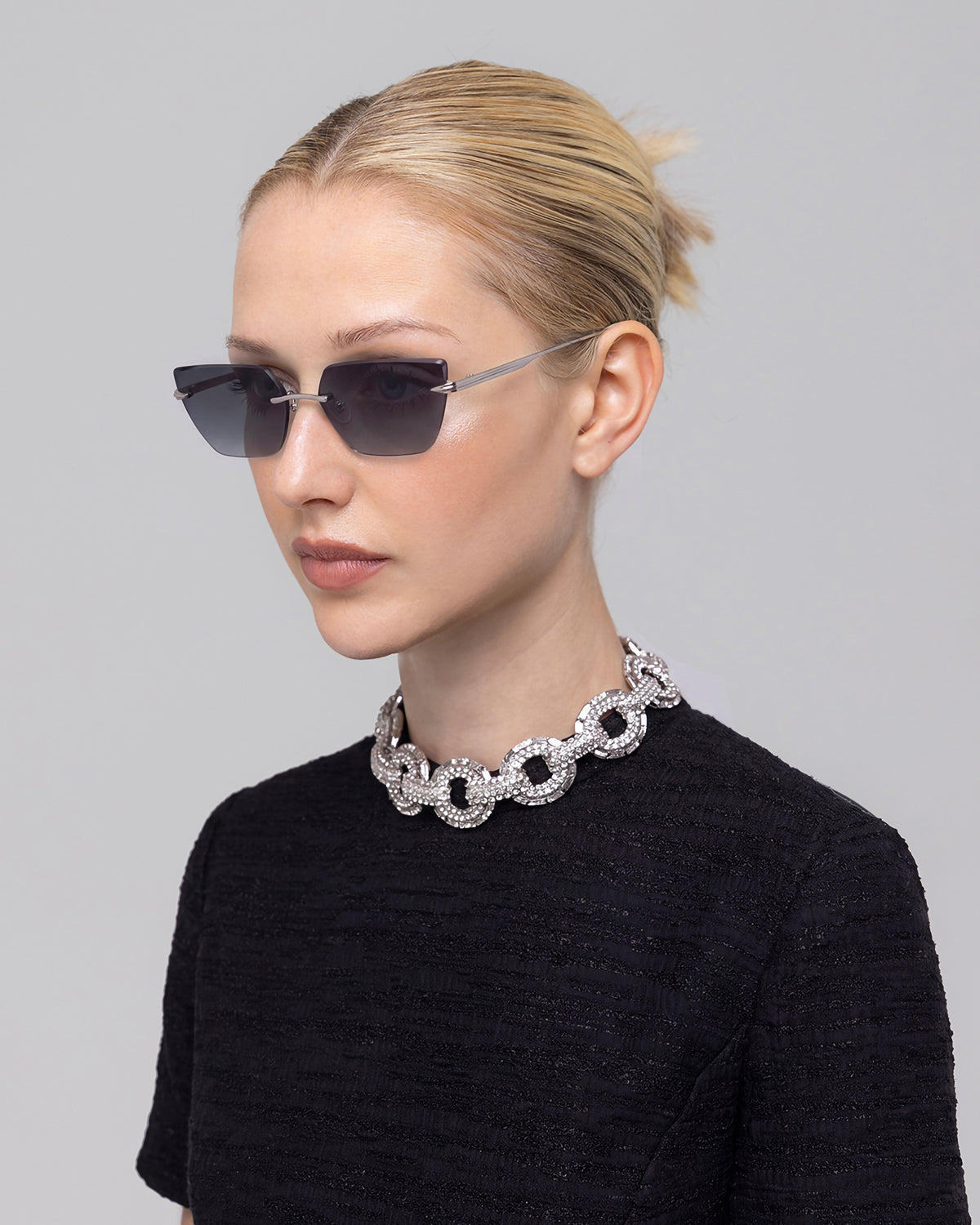 Blonde woman in a bun wears For Art’s Sake® Sienna II sunglasses with jade stone nose pads, a textured black top, and a bold silver chain necklace against a plain light gray background.