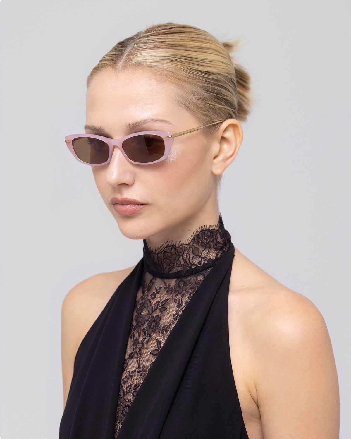 A woman with blonde hair in a low bun wears For Art’s Sake® Marisol I sunglasses and a black lace halter top, looking slightly to the side against a plain light background.