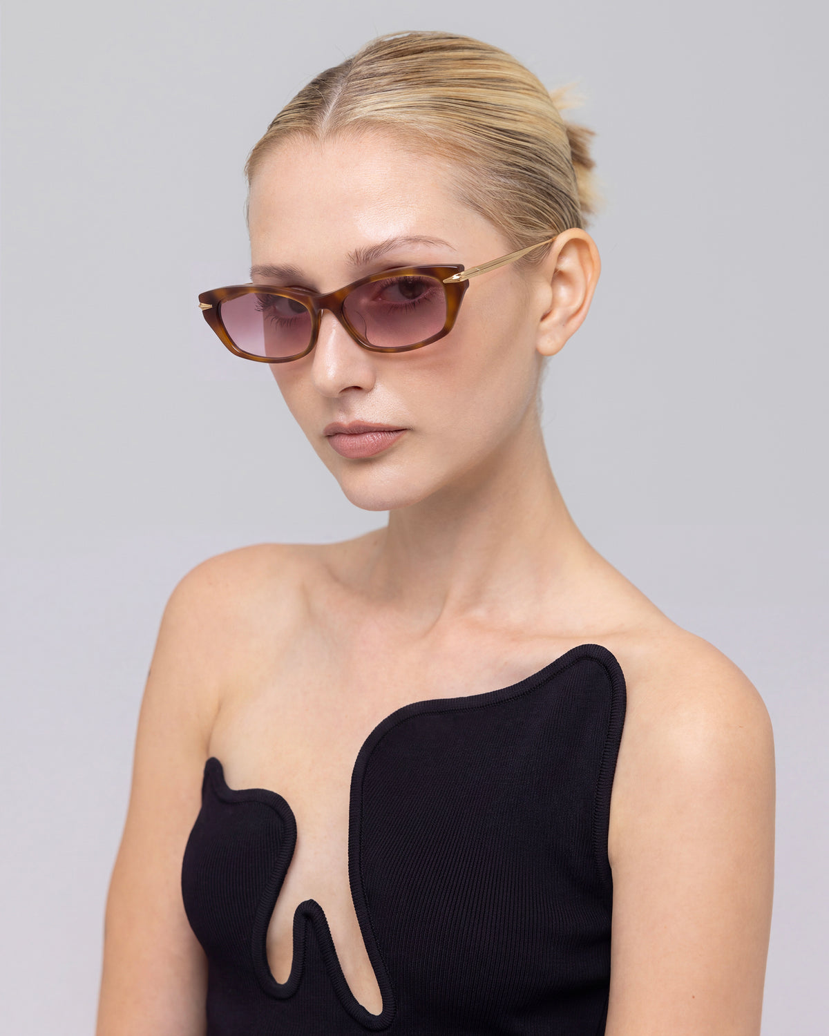 A woman with slicked-back blonde hair wears Marisol I by For Art’s Sake® and a black top with a wavy neckline, posing against a gray background for effortless everyday sun protection.