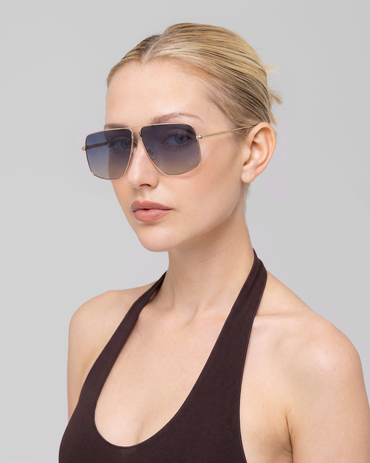 A woman with blonde hair in a neat bun wears For Art’s Sake® Marisol II oversized aviator sunglasses with sculpted metal detailing and a dark halter top, facing slightly to the side against a light gray background.