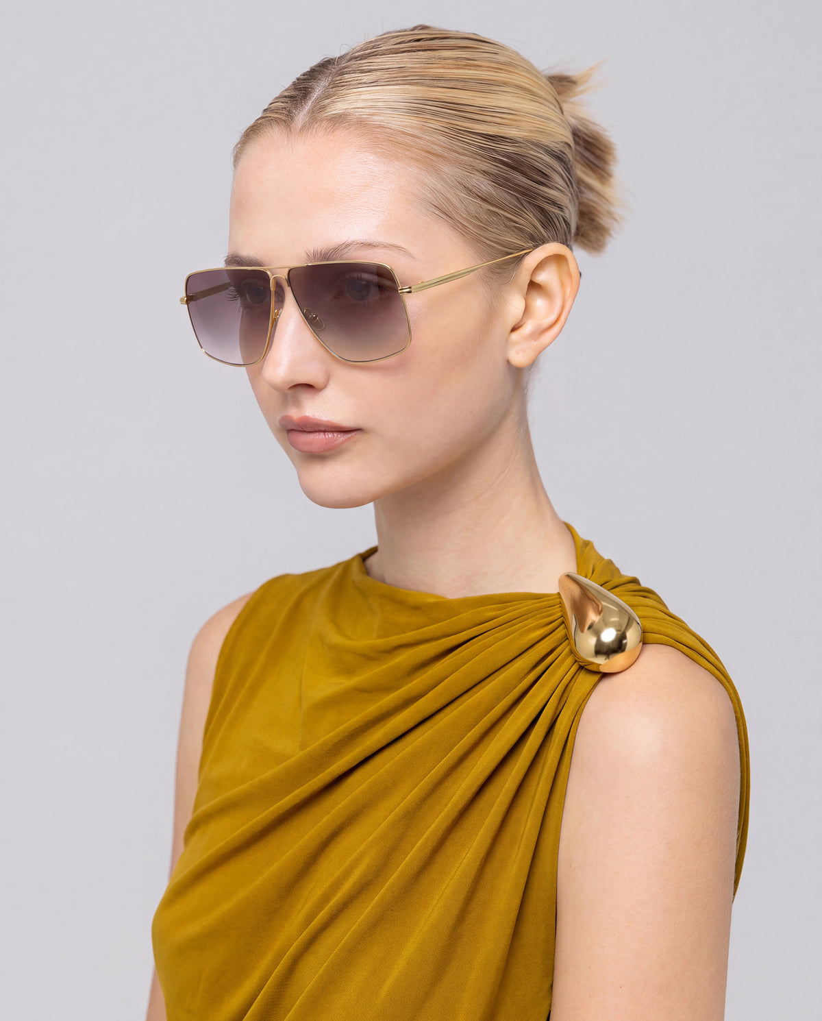 A woman with blond hair in a bun wears oversized For Art’s Sake® Marisol II aviator sunglasses and a mustard-yellow dress, posing against a plain light background.