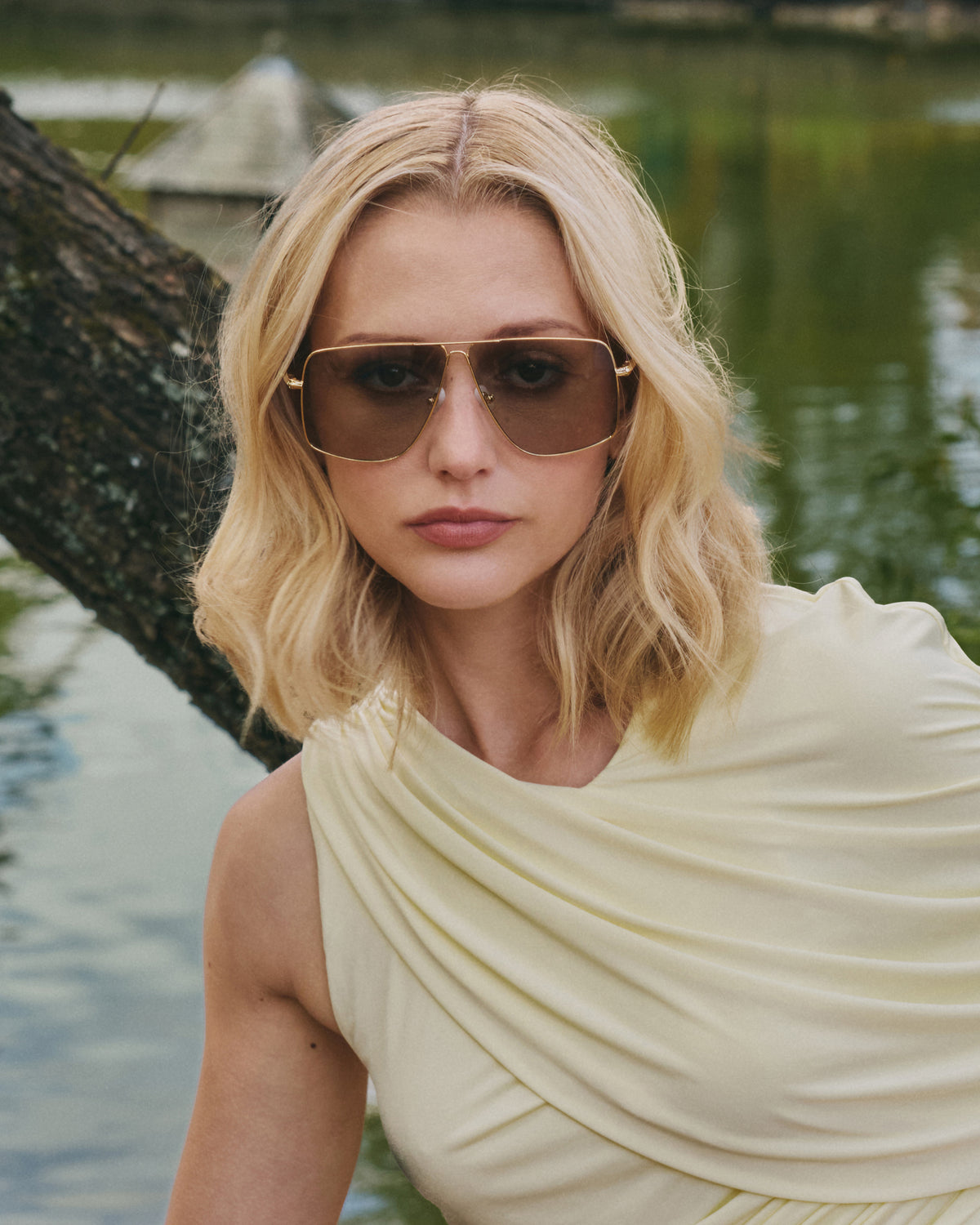 A woman with wavy blonde hair wears Marisol II by For Art’s Sake®—oversized aviator sunglasses with sculpted metal details—posing outdoors in a light cream sleeveless top near a tree and pond.
