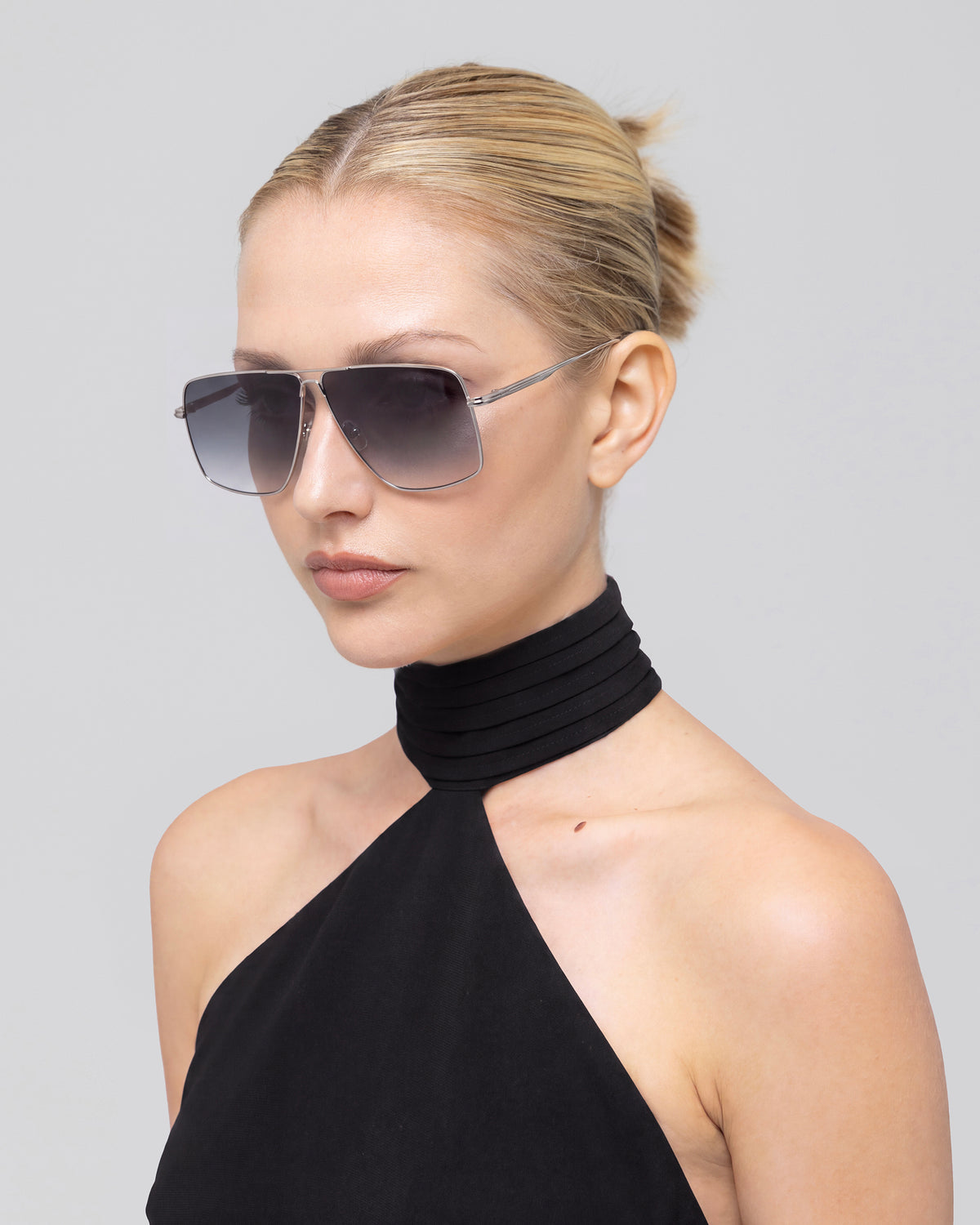 A woman with blonde hair in a bun wears For Art’s Sake® Marisol II oversized aviator sunglasses and a black halter neck top, posing against a plain light gray background.