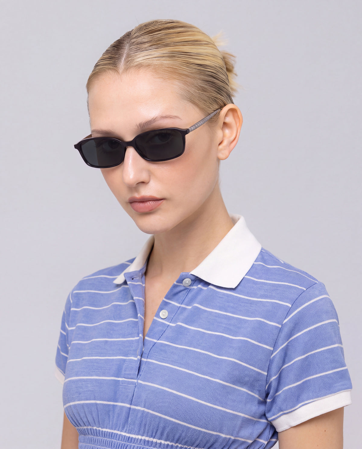 A woman with blond hair in a bun wears For Art’s Sake® Solana I bold black sunglasses and a blue collared shirt with white stripes, standing against a plain light background.