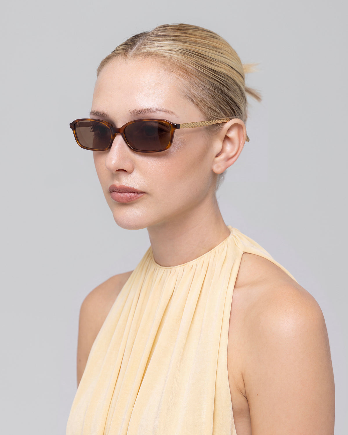 A woman with blonde hair in a low bun wears For Art’s Sake® Solana I brown rectangular sunglasses and a sleeveless light beige pleated top, gazing slightly to the side against a plain light gray background.