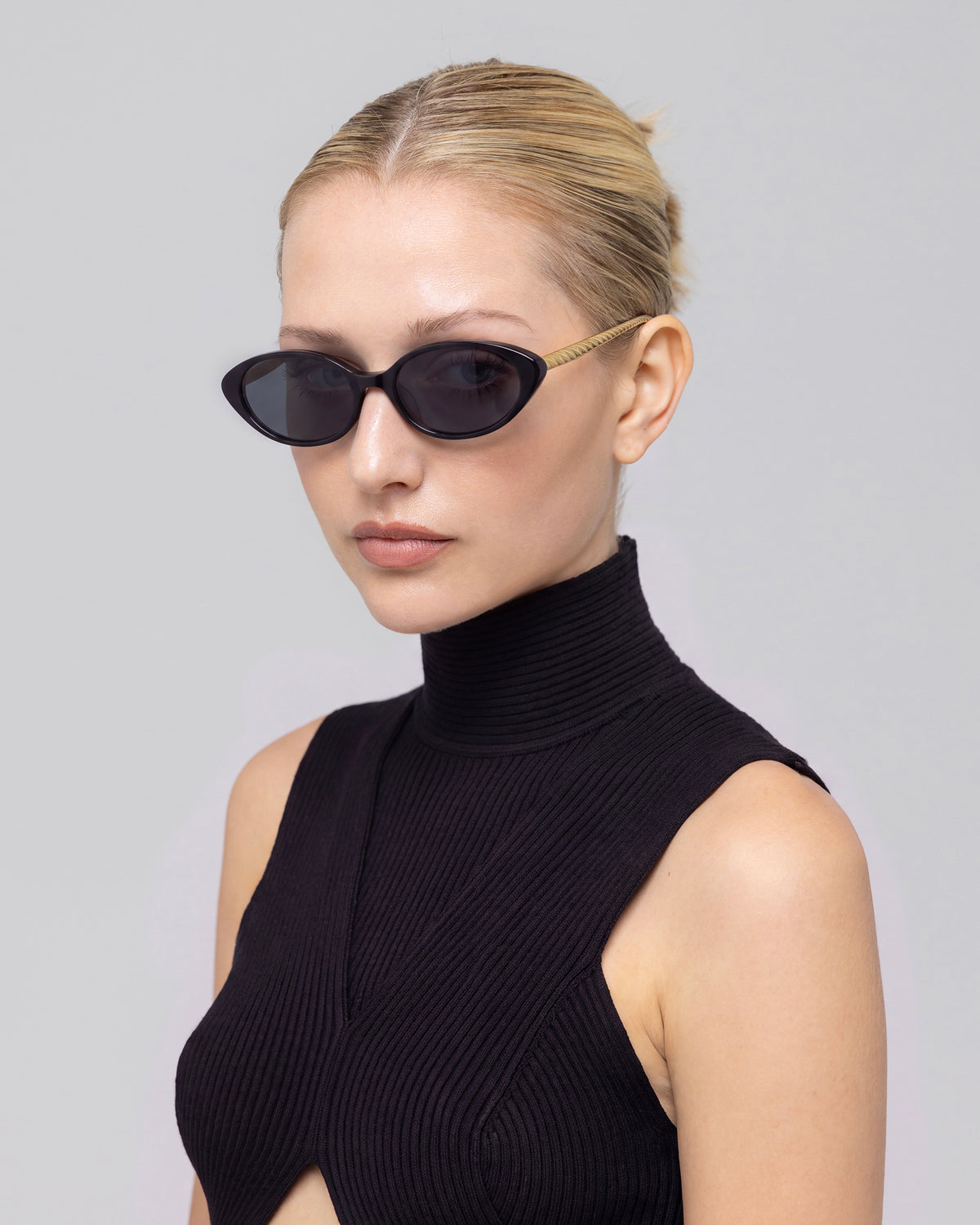 A woman with blond hair in a low bun wears For Art’s Sake® Solana II black oval acetate sunglasses and a sleeveless black turtleneck top with a front cutout, posing against a plain light background.
