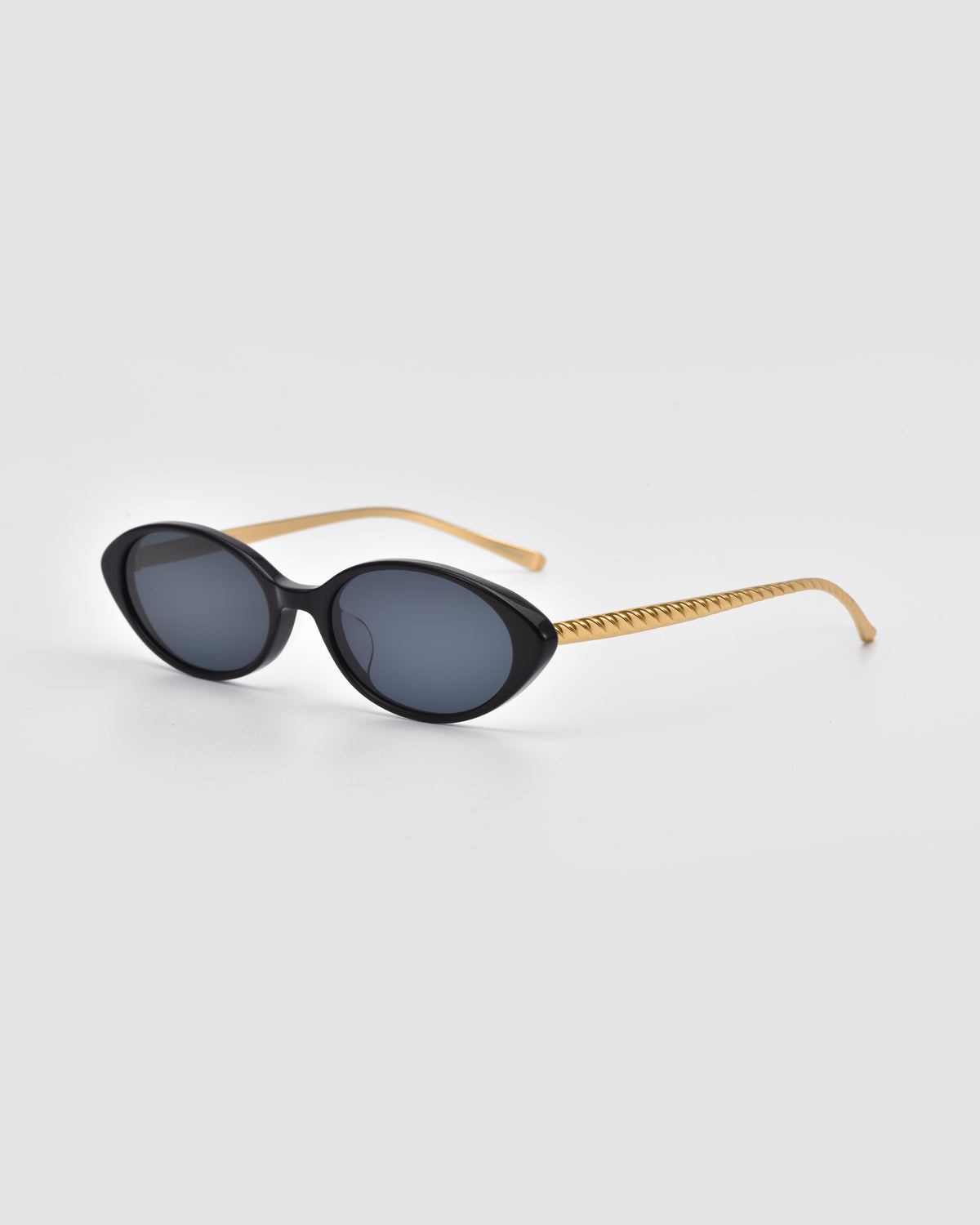 Solana II by For Art’s Sake®: black oval acetate sunglasses with dark lenses and gold-tone textured arms, displayed on a light gray background for a striking statement look.