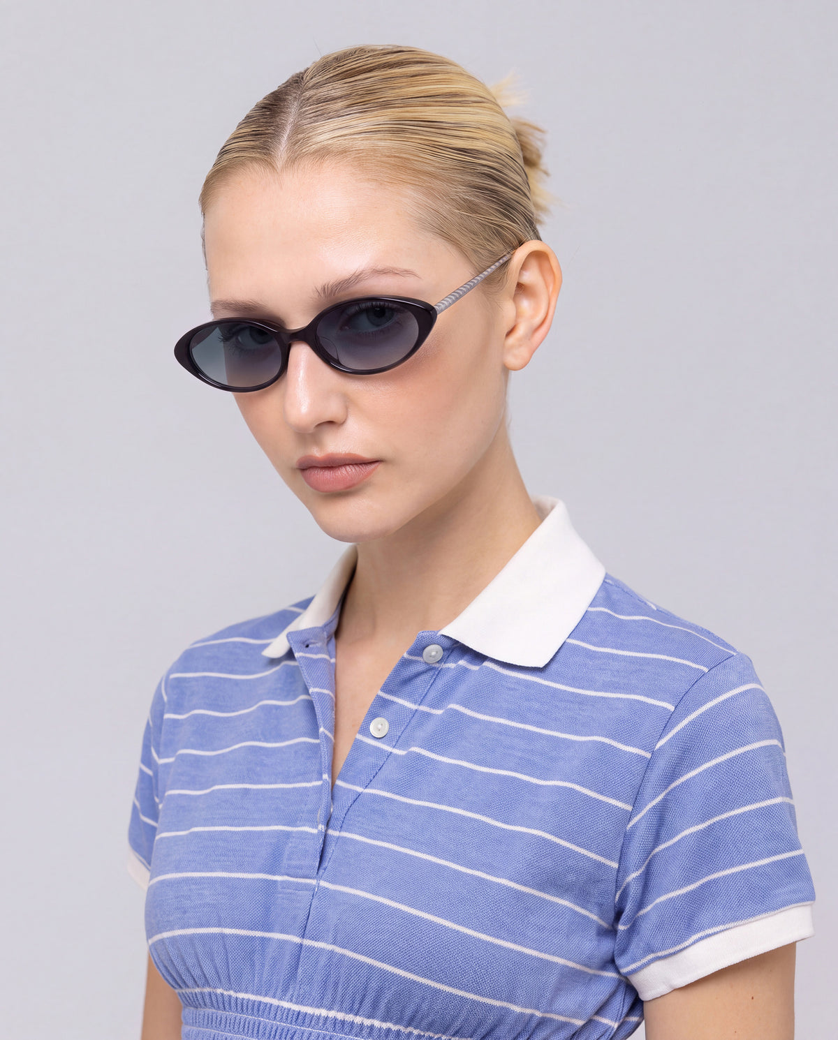 Woman with blond hair tied back wears For Art’s Sake® Solana II dark oval acetate sunglasses and a blue collared shirt with white stripes against a light background for a chic statement look.