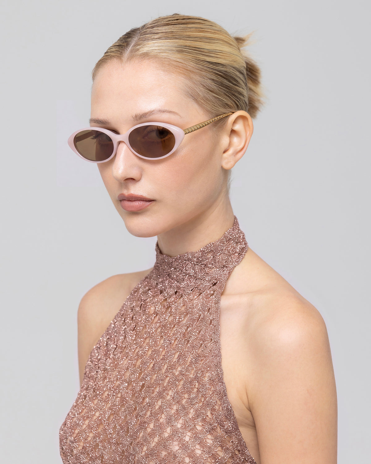 A woman with blonde hair in a bun wears For Art’s Sake® Solana II pink oval sunglasses and a shimmery, sleeveless high-neck top, gazing to the side against a plain gray background.