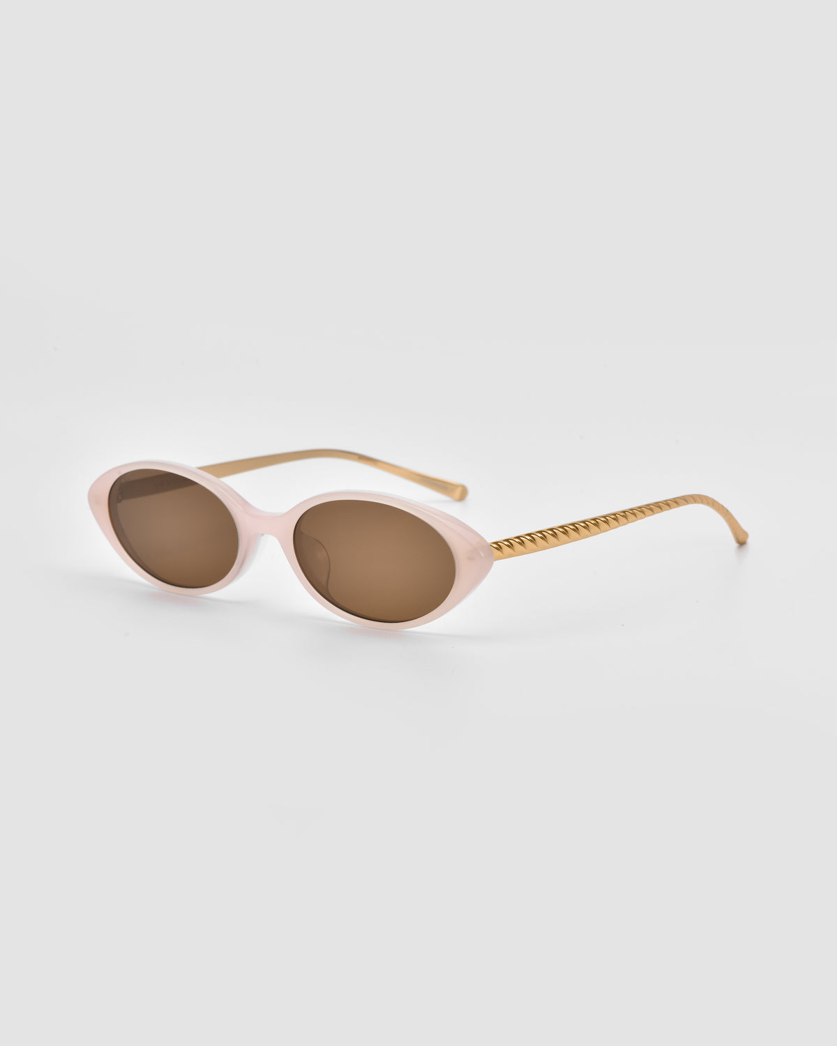 Solana II by For Art’s Sake®: Oval acetate sunglasses with light pink frames, brown lenses, and gold textured arms shown on a light gray background.