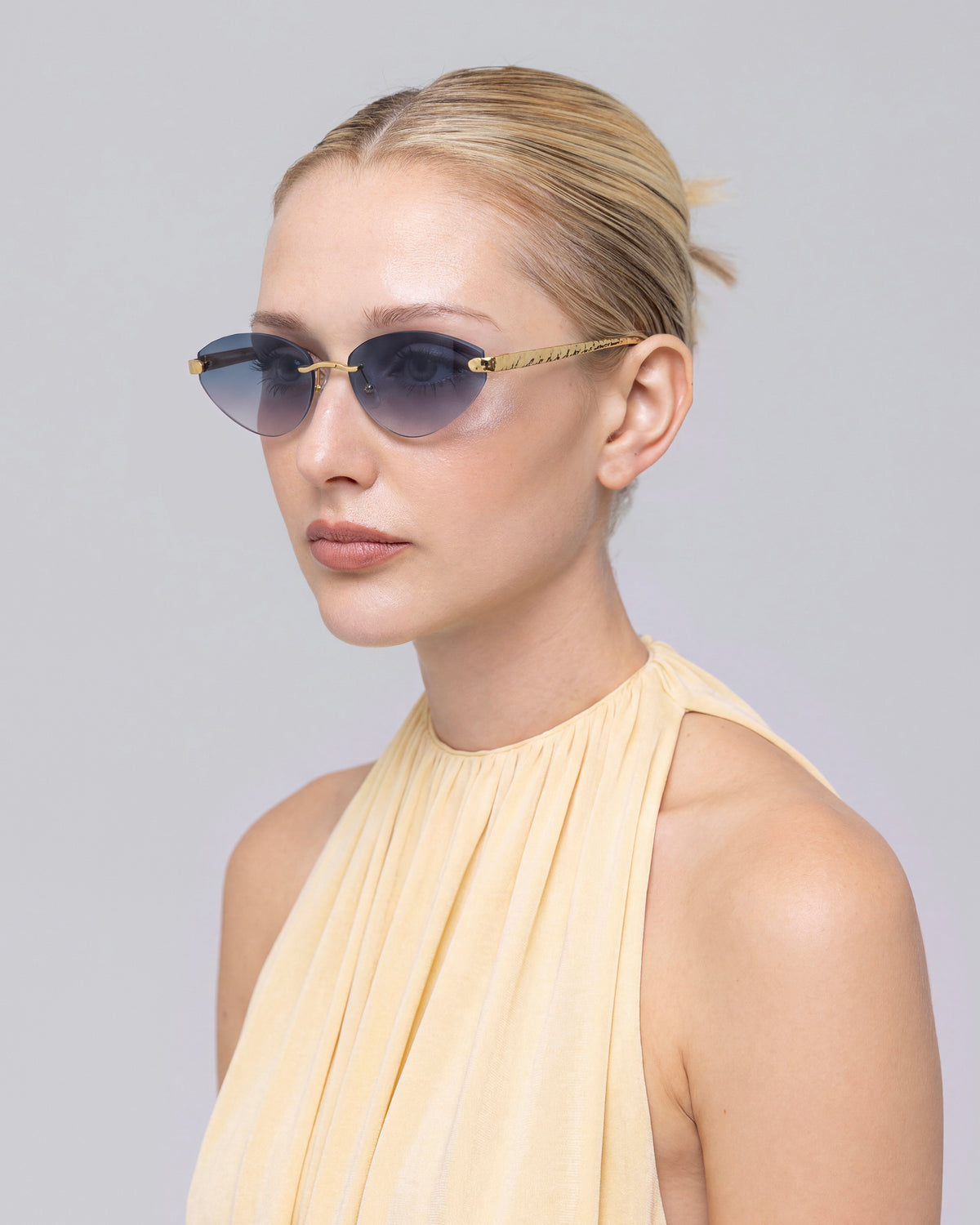A woman with blonde hair tied back wears Shimmer I sunglasses by For Art’s Sake® and a sleeveless, light beige top, looking forward against a neutral background.
