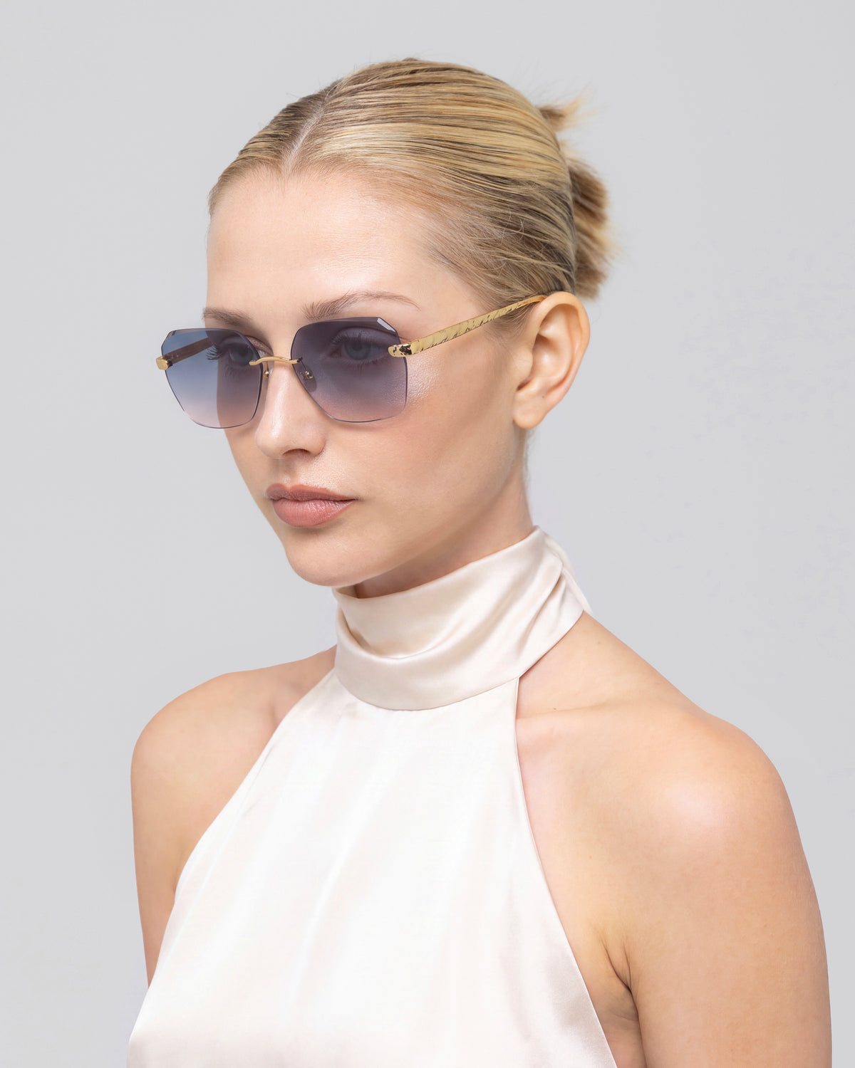A blonde woman with her hair in a bun wears For Art’s Sake® Shimmer II sunglasses and a sleeveless cream satin top, posing against a plain gray background.