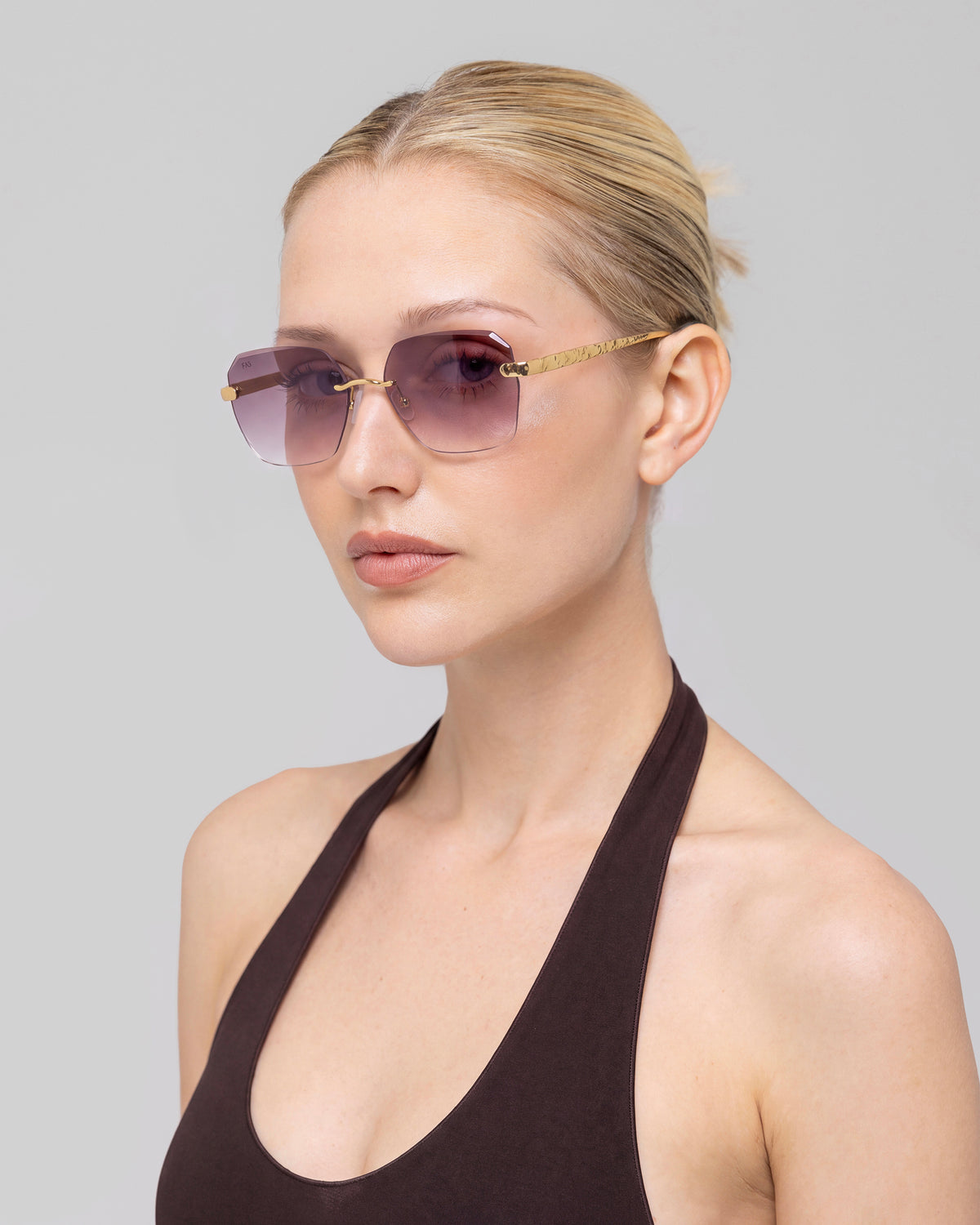 A blonde-haired woman in a bun wears For Art’s Sake® Shimmer II rimless sunglasses and a dark halter top, posing against a plain light gray background.