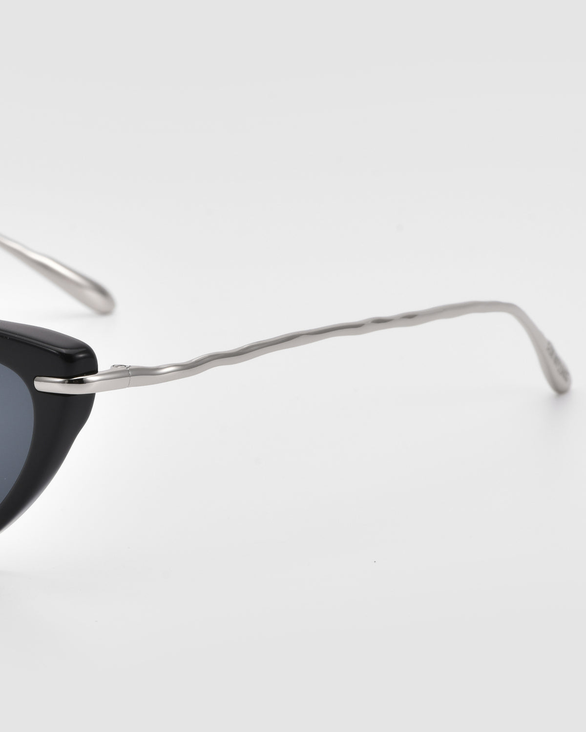 Close-up of Aurora cat-eye sunglasses by For Art’s Sake® featuring a black frame and slim, wavy jewellery-inspired sculpted metal temples against a plain light gray background.