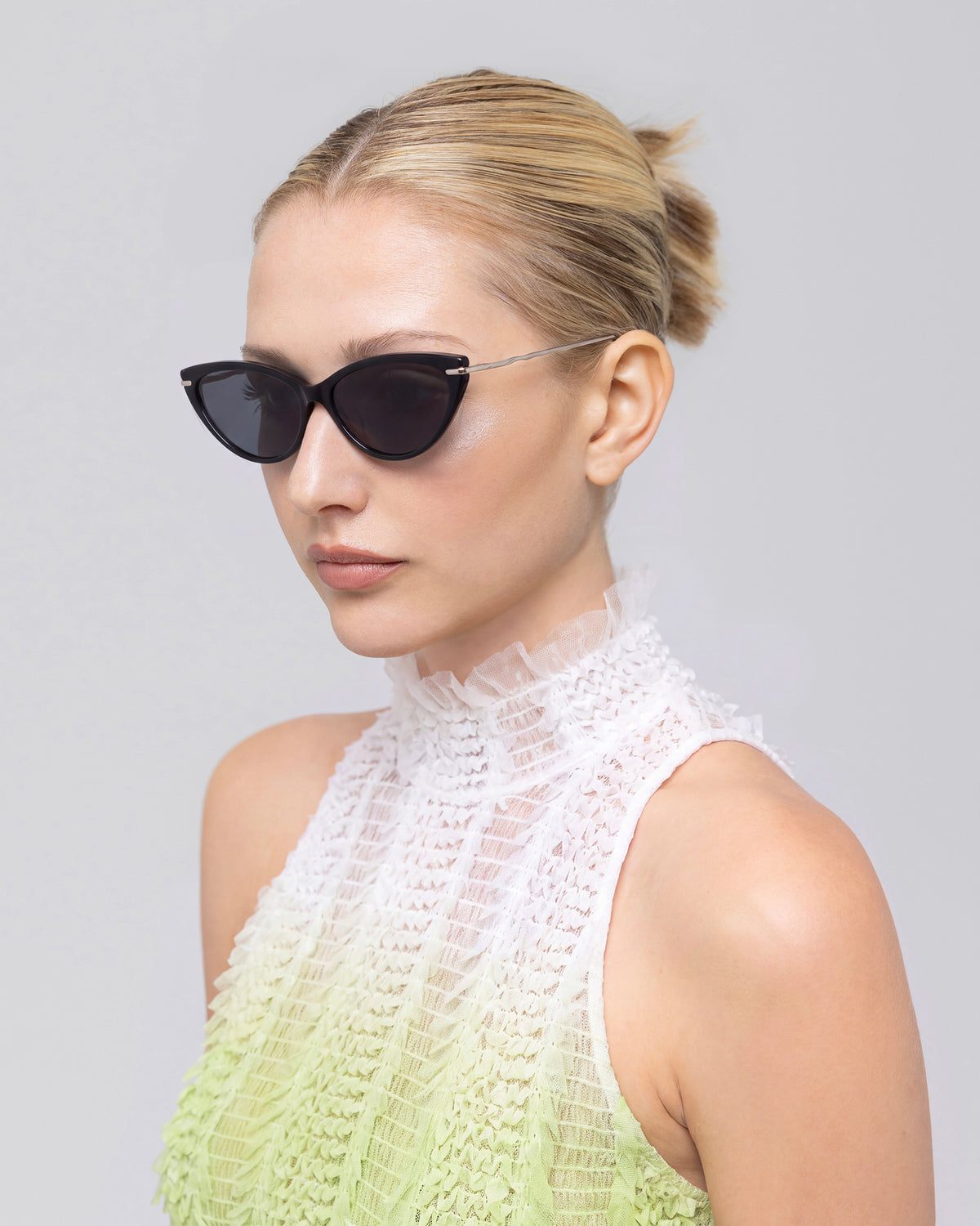 A woman with blonde hair in a neat bun wears For Art’s Sake® Aurora black cat-eye sunglasses with sculpted metal temples and a white textured high-neck sleeveless top, looking to the side against a plain background.