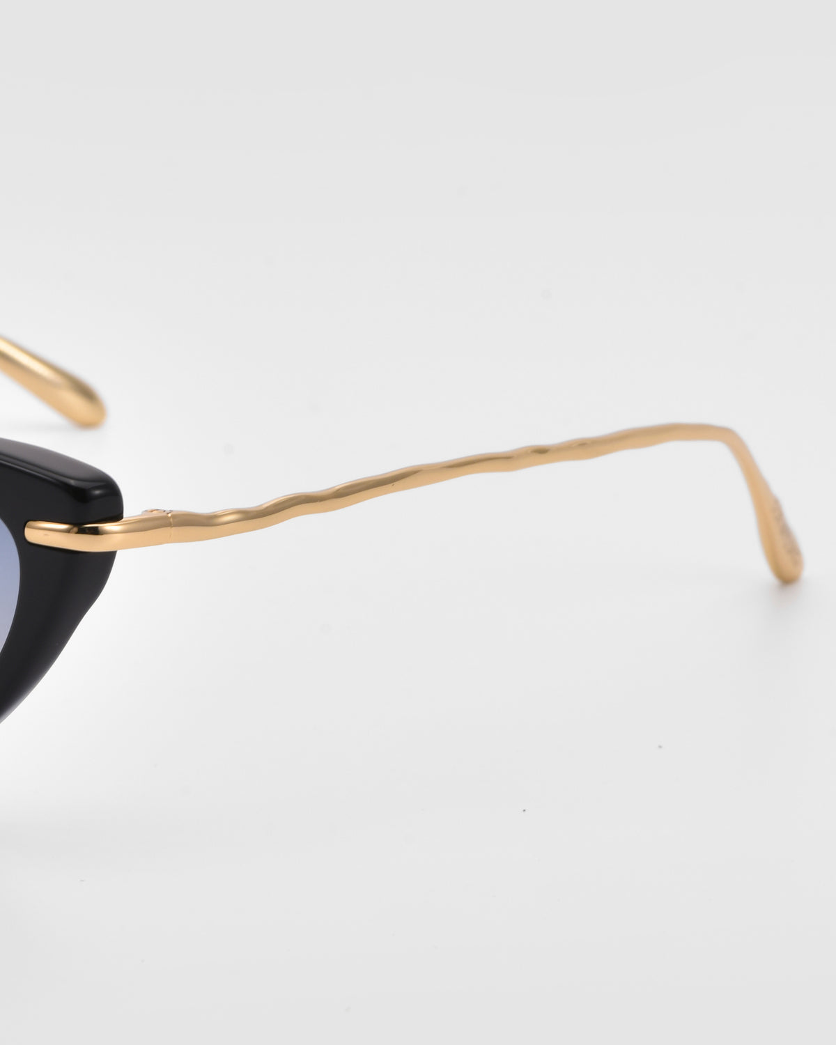 Close-up of For Art’s Sake® Aurora sunglasses featuring a black cat-eye silhouette and slim, sculpted gold metal temples on a plain white background.