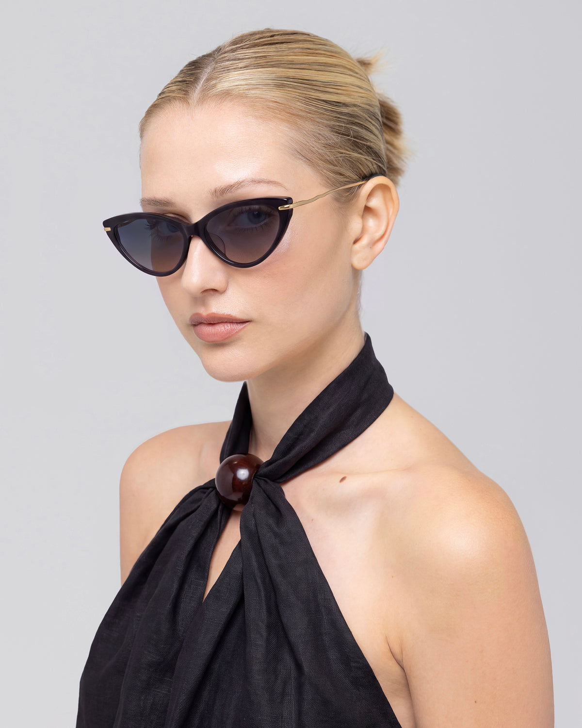 A blonde woman with a bun wears For Art’s Sake® Aurora black cat-eye sunglasses with sculpted metal temples and a black halter top, posing against a plain light gray background.