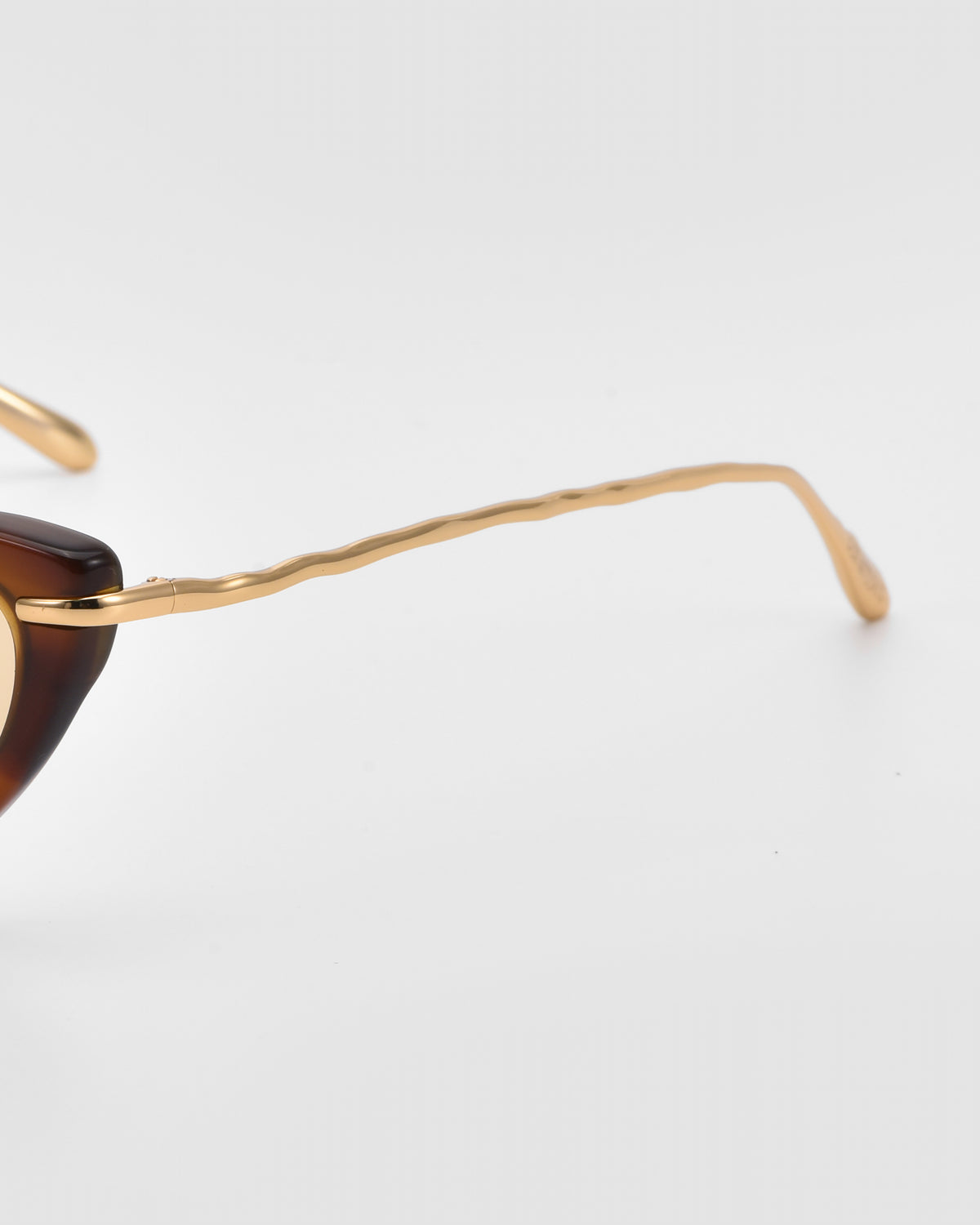 Close-up of For Art’s Sake® Aurora sunglasses with a tortoiseshell frame and gold sculpted metal temples, set on a plain light gray background.
