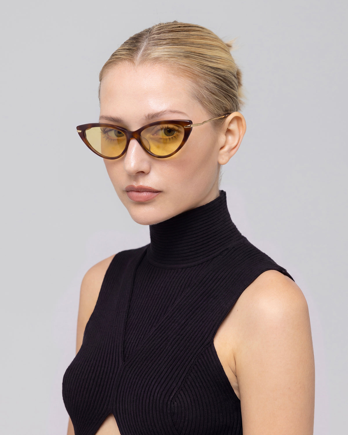 A woman with blonde hair in a bun wears For Art’s Sake® Aurora sunglasses and a black sleeveless turtleneck, posing against a plain light gray background.