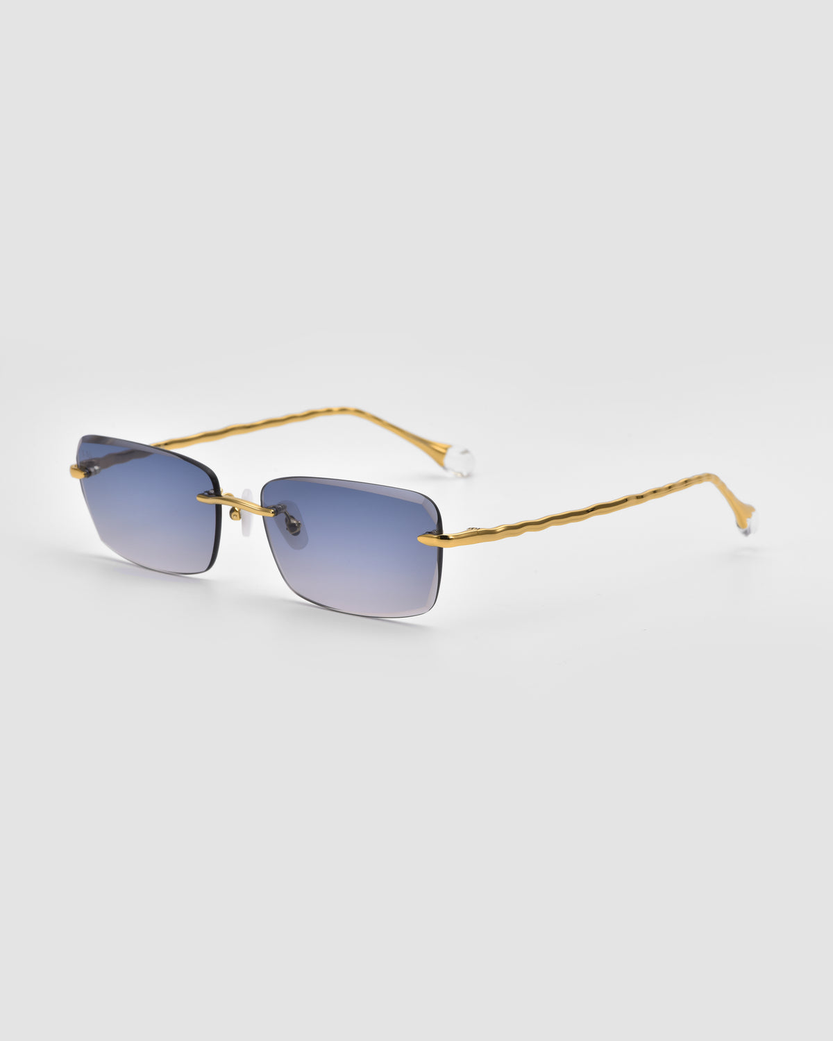 Sol I sunglasses by For Art’s Sake® feature gold-rimmed translucent jewel frames, square blue-tinted lenses, and wavy gold arms with sculpted nylon tips, shown against a light gray background.